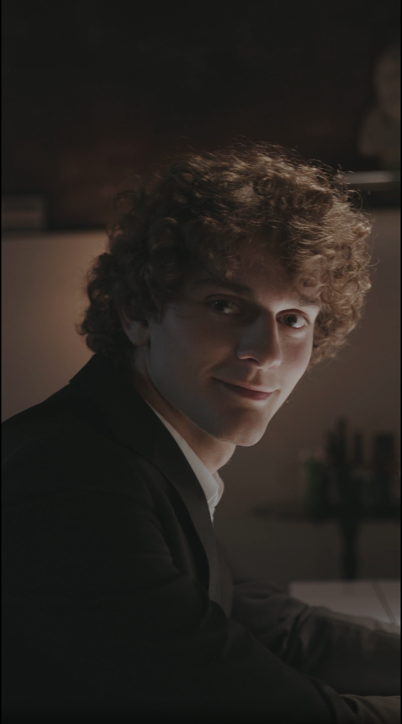 Curly-haired guy looking at the camera across a dim room